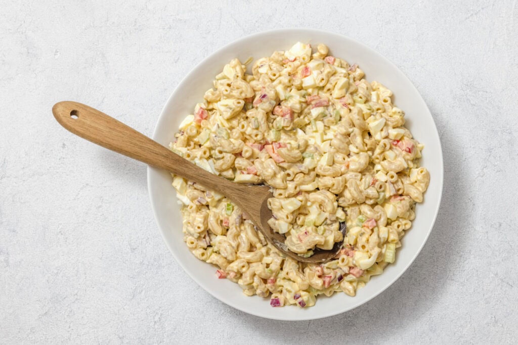 A bowl of macaroni salad with diced vegetables, including red peppers and celery, and a wooden spoon resting on top.
