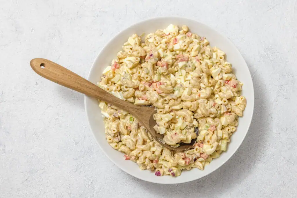 A bowl of macaroni salad with diced vegetables, including red peppers and celery, and a wooden spoon resting on top.
