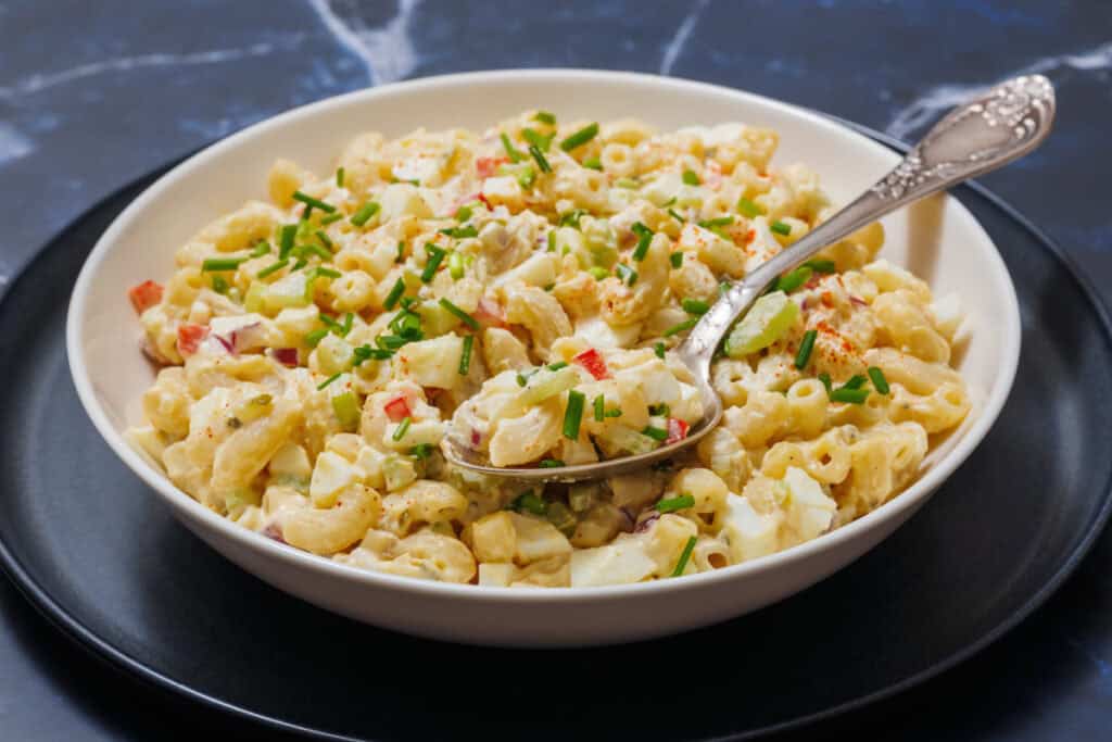 A bowl of Deviled Egg Pasta Salad with chopped vegetables and herbs, is set on a black plate, and a spoon rests in it.