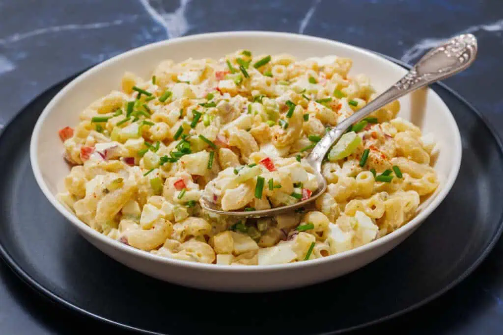 A bowl of Deviled Egg Pasta Salad with diced vegetables and herbs, served with a spoon on a dark plate.