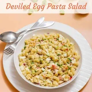 A bowl of deviled egg pasta salad with macaroni, diced vegetables, and herbs. A spoon and fork are placed on the side.