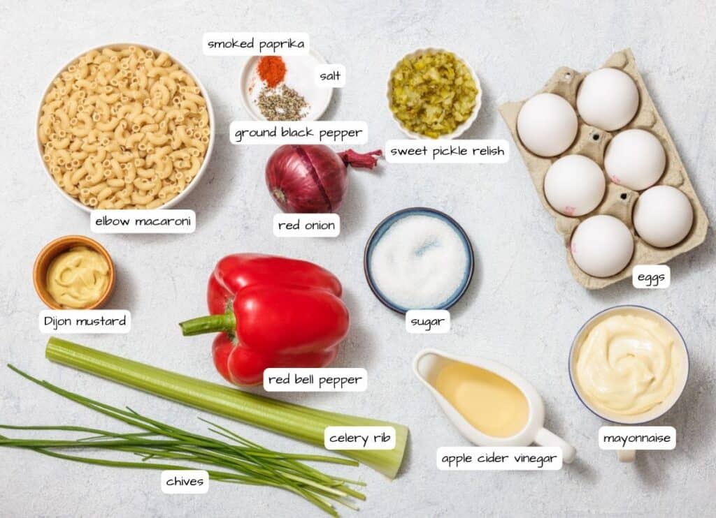 Labeled ingredients for a Deviled Egg Pasta Salad recipe, including elbow macaroni, eggs, mayonnaise, red bell pepper, celery, chives, and spices, arranged on a light surface.