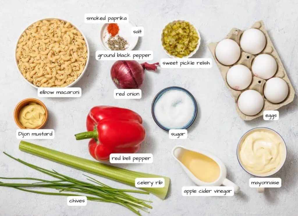 Labeled ingredients for a Deviled Egg Pasta Salad recipe, including elbow macaroni, eggs, mayonnaise, red bell pepper, celery, chives, and spices, arranged on a light surface.