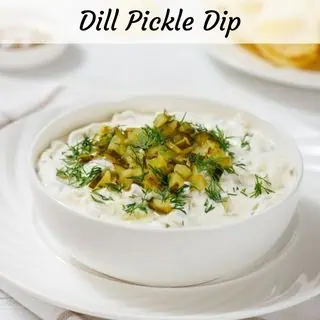 A bowl of dill pickle dip garnished with chopped pickles and dill, set on a white plate.