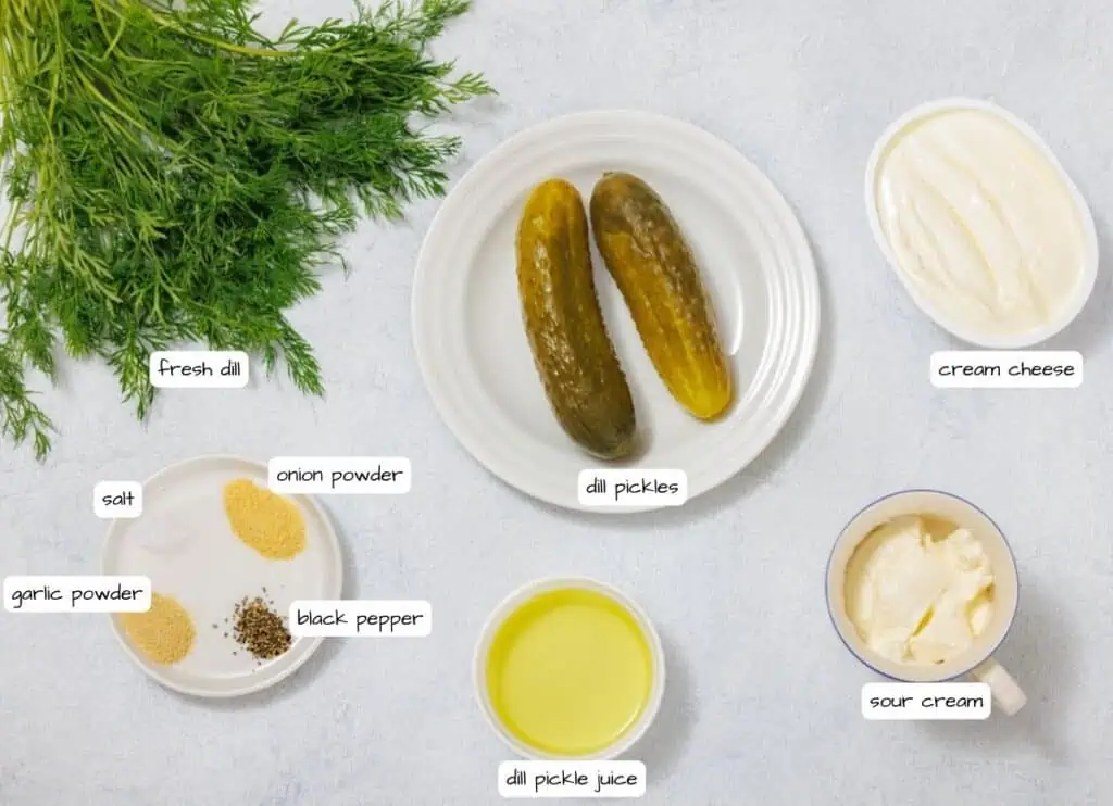Ingredients for a dill pickle dip: fresh dill, dill pickles, pickle juice, cream cheese, sour cream, salt, black pepper, onion powder, garlic powder, arranged on a light surface.