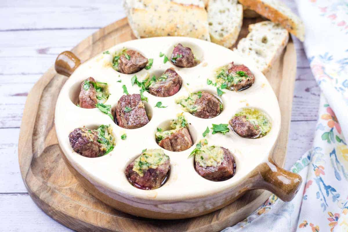 Garlic Butter Steak Bites Escargot-Style dish in a round ceramic plate with cooked snails topped with herbs and butter sauce, and slices of bread on a wooden board.