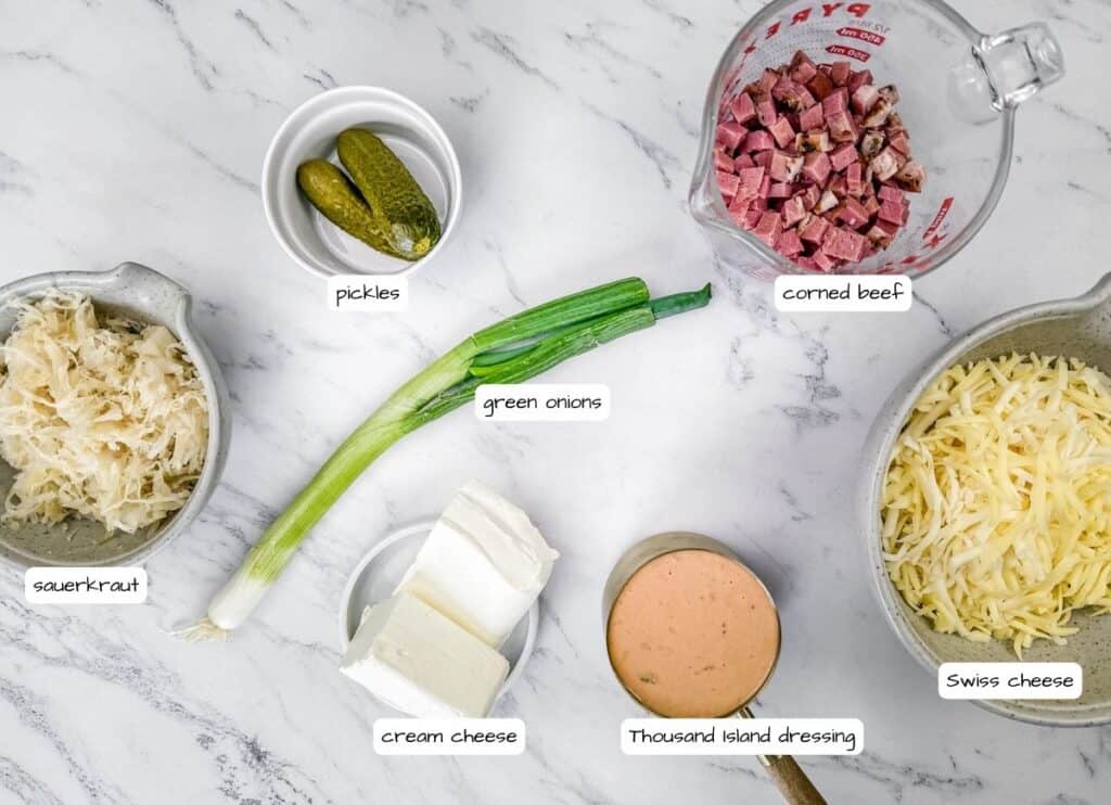 Ingredients for a Reuben dip arranged on a marble surface include sauerkraut, pickles, green onions, cream cheese, corned beef, Thousand Island dressing, and Swiss cheese.