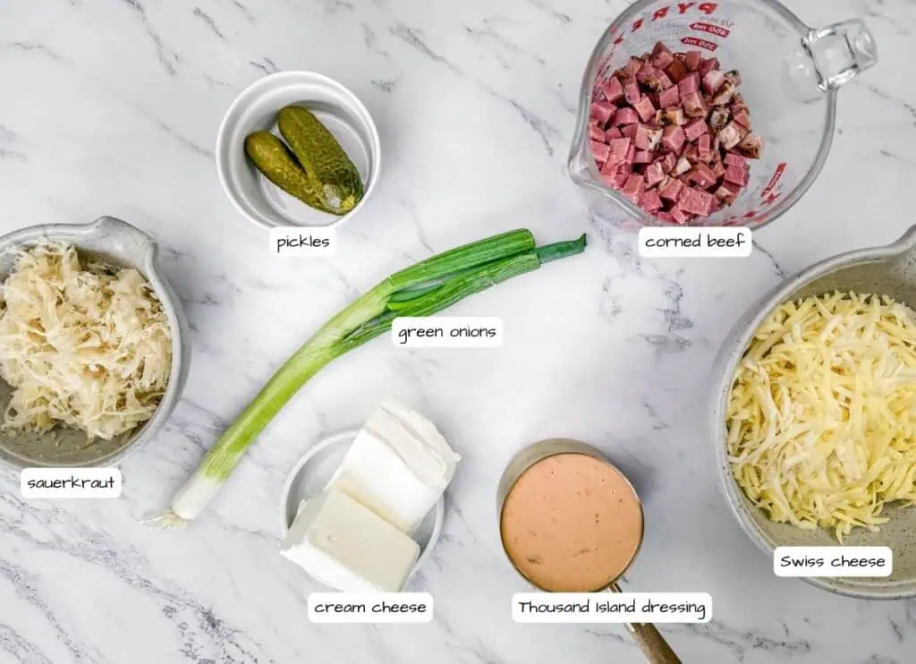 Ingredients for a Reuben dip arranged on a marble surface include sauerkraut, pickles, green onions, cream cheese, corned beef, Thousand Island dressing, and Swiss cheese.