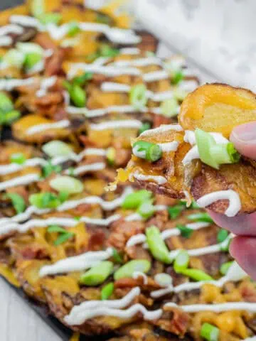 Hand holding a loaded Irish nachos with cheese, bacon, sour cream, and green onions, over a platter of similarly topped slices on a table.
