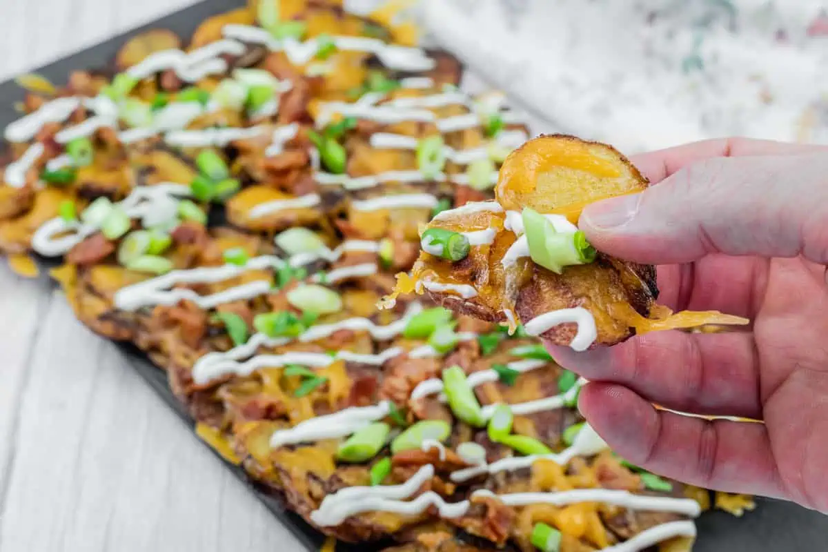 Hand holding a loaded Irish nachos with cheese, bacon, sour cream, and green onions, over a platter of similarly topped slices on a table.