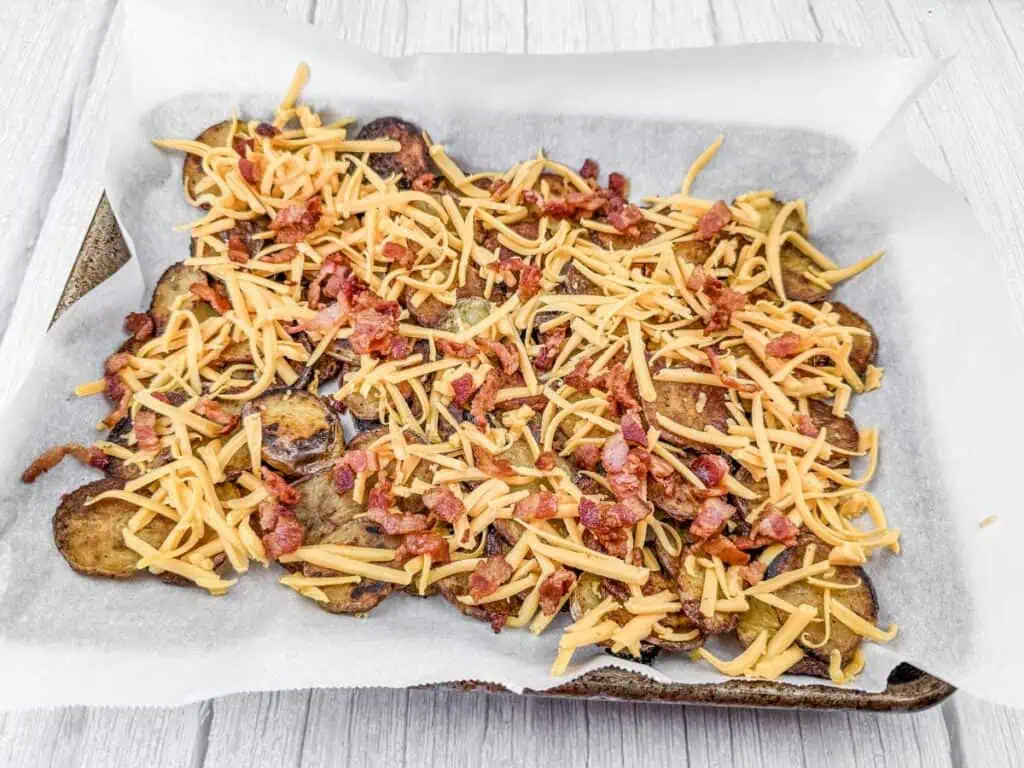 Baked potato slices on a parchment-lined tray, topped with shredded cheese and bacon bits.