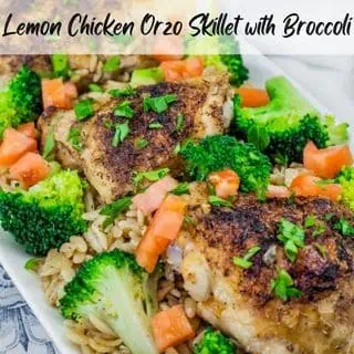 A Lemon Chicken Orzo Skillet with Broccoli dish featuring golden-brown seasoned chicken thighs on a bed of orzo pasta, garnished with fresh broccoli florets, diced tomatoes, chopped parsley, and lemon slices in the background.