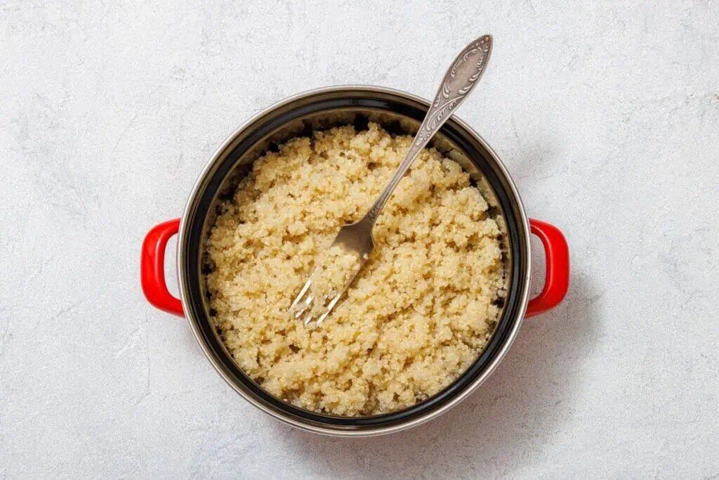 A pot with red handles filled with cooked quinoa, with a metal fork resting inside on a light gray background.