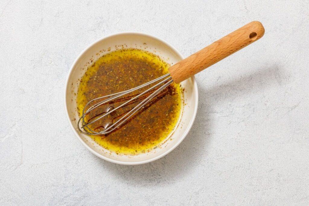 Bowl containing a mixture of oil and spices with a whisk resting inside, placed on a white surface.