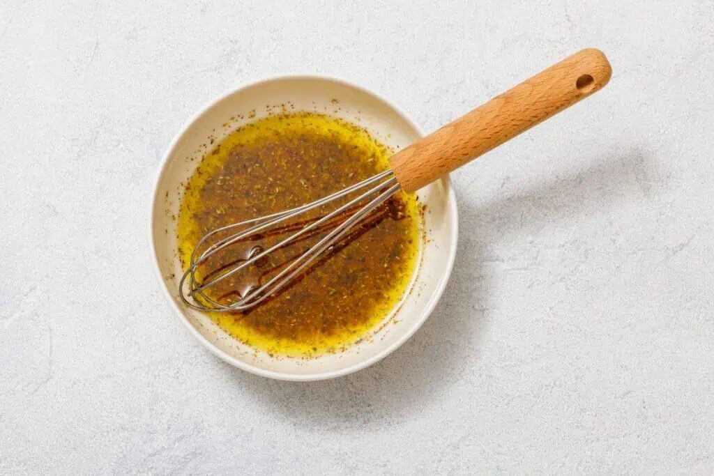 Bowl containing a mixture of oil and spices with a whisk resting inside, placed on a white surface.