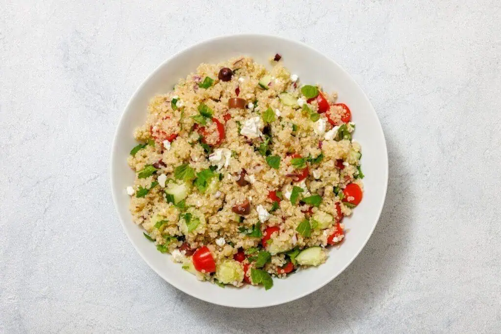 A bowl of quinoa salad with cherry tomatoes, cucumber, olives, feta cheese, and fresh herbs on a light textured surface.