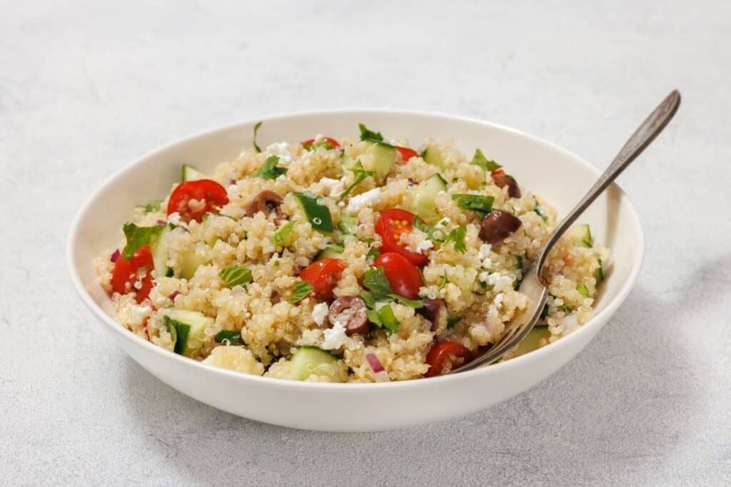 A bowl of quinoa salad with cherry tomatoes, cucumbers, olives, feta cheese, and herbs. A spoon rests inside the bowl on a light surface.