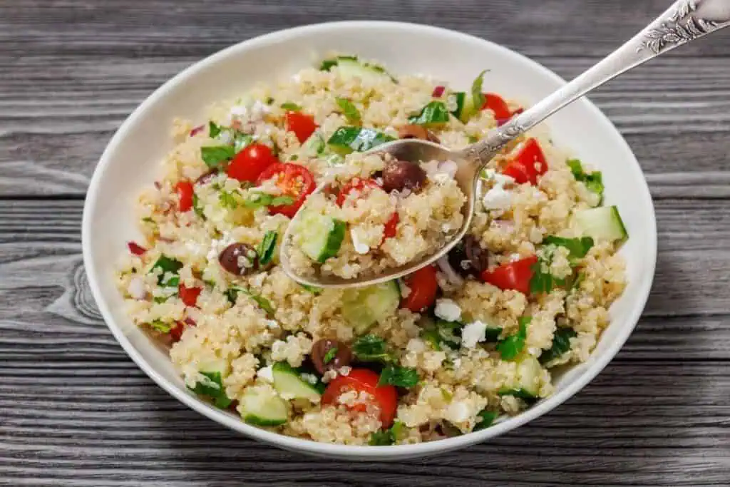 A bowl of quinoa salad with cherry tomatoes, cucumbers, olives, feta cheese, and herbs, with a spoon resting inside. The bowl is on a wooden surface.