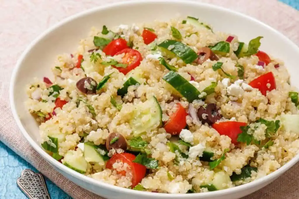 A bowl of Mediterranean Quinoa Salad with cherry tomatoes, cucumber, olives, red onion, feta cheese, and parsley on a light blue tablecloth.
