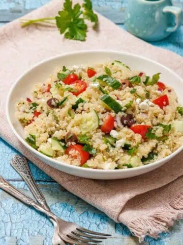 A bowl of quinoa salad with cucumbers, tomatoes, olives, and feta on a pink cloth. A small jug in the background and sprig of parsley on light blue wooden surface.