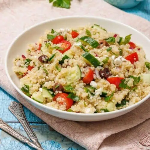 A bowl of quinoa salad with cucumbers, tomatoes, olives, and feta on a pink cloth. A small jug in the background and sprig of parsley on light blue wooden surface.
