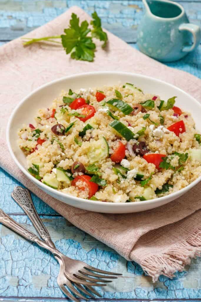 A bowl of quinoa salad with cucumbers, tomatoes, olives, and feta on a pink cloth. A small jug in the background and sprig of parsley on light blue wooden surface.