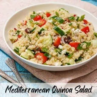A bowl of Mediterranean quinoa salad with cucumber, tomatoes, olives, and herbs, placed on a cloth beside two forks.