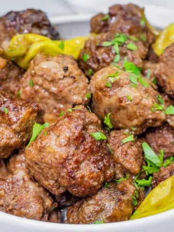 Bowl of browned Slow Cooker Mississippi Meatballs garnished with chopped parsley and yellow pepperoncini peppers.