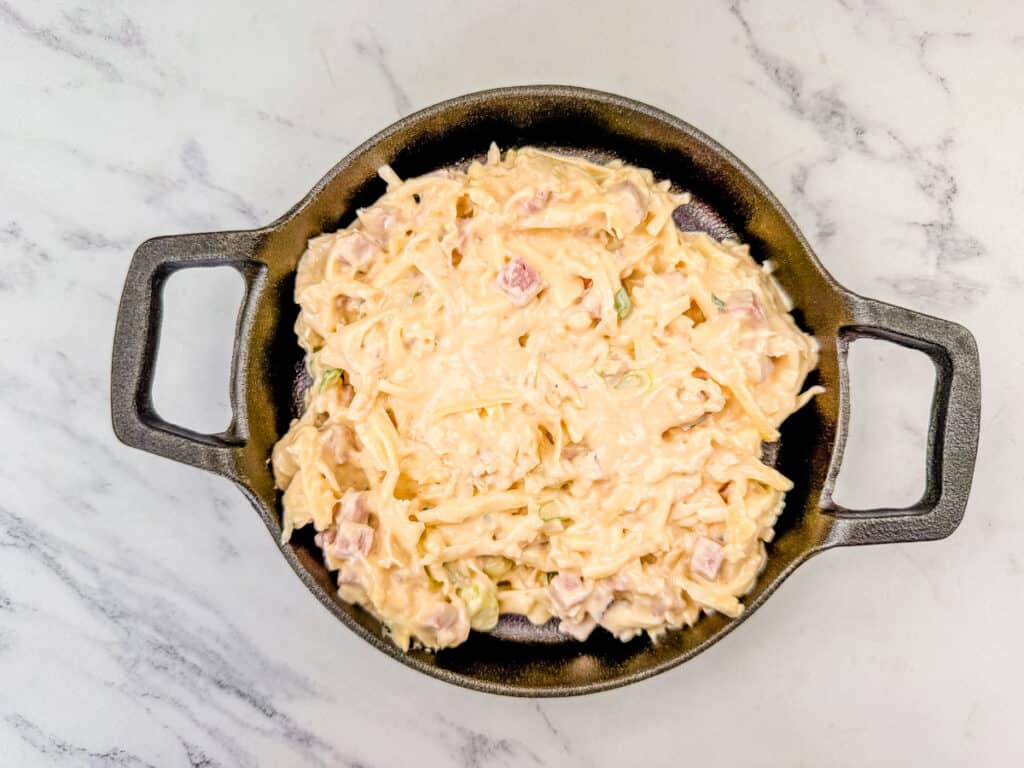 Top view of a creamy dish with diced ham and shredded cheese in a round black pan on a marble surface.