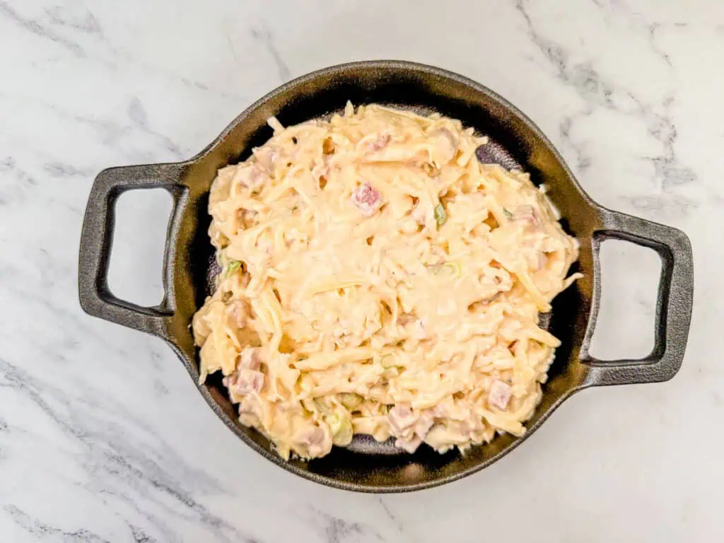 Top view of a creamy dish with diced ham and shredded cheese in a round black pan on a marble surface.