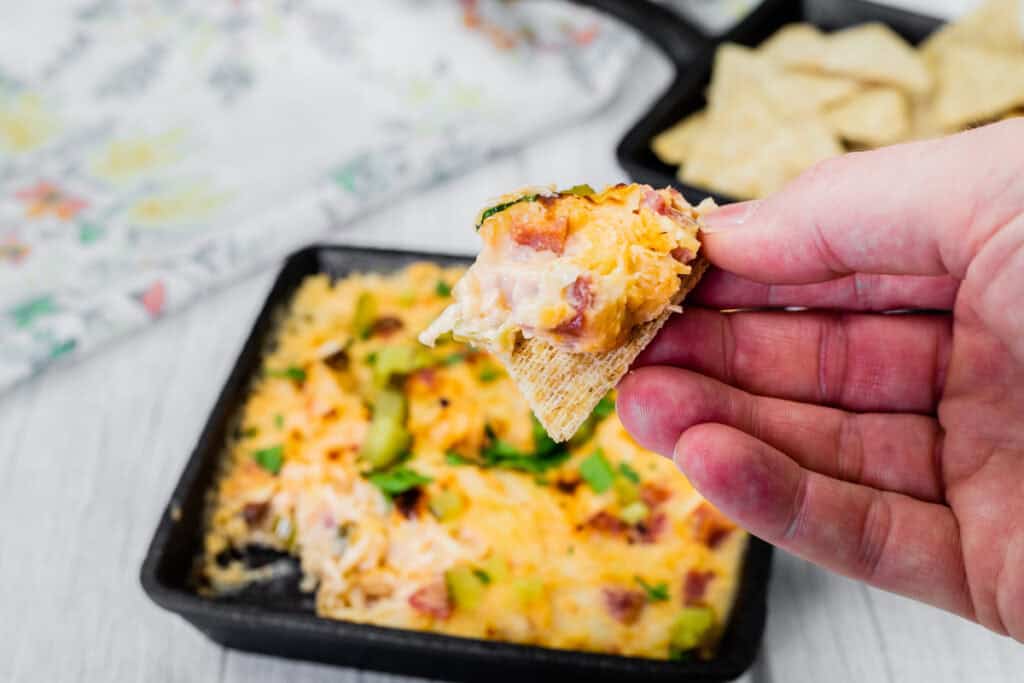 A hand holds a chip with Hot Reuben Dip from a skillet of the dip and cracker.