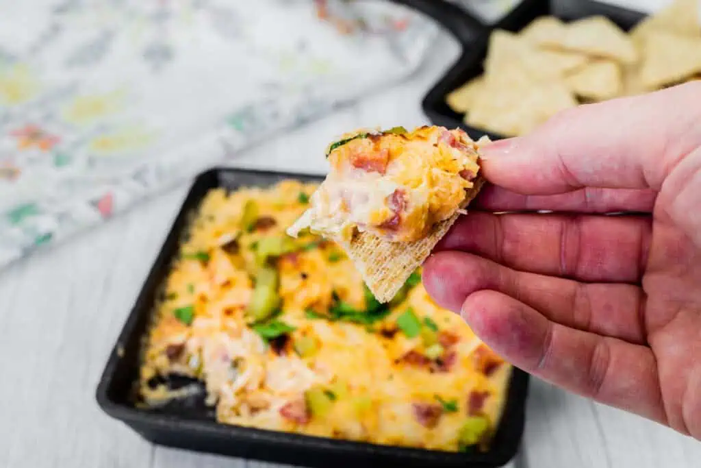 A hand holds a chip with Hot Reuben Dip from a skillet of the dip and cracker.