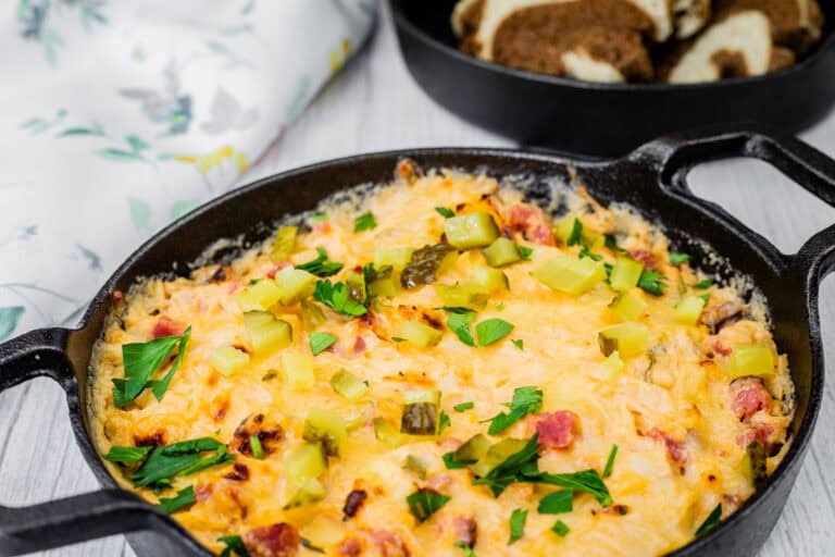 Cheesy, golden-brown Hot Reuben dip in a cast-iron skillet, topped with chopped pickles and fresh parsley, served with slices of marbled rye bread.