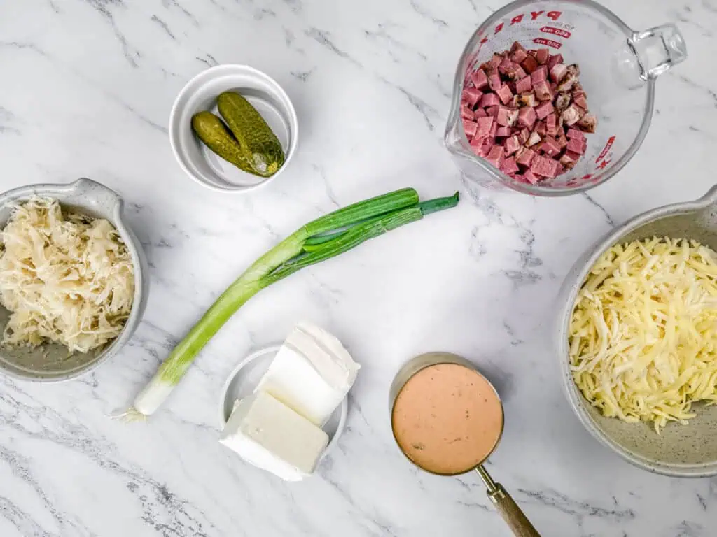 Ingredients on a marble surface: sauerkraut, pickles, chopped meat in a measuring cup, shredded cheese, cream cheese, sliced green onion, and a cup of sauce.