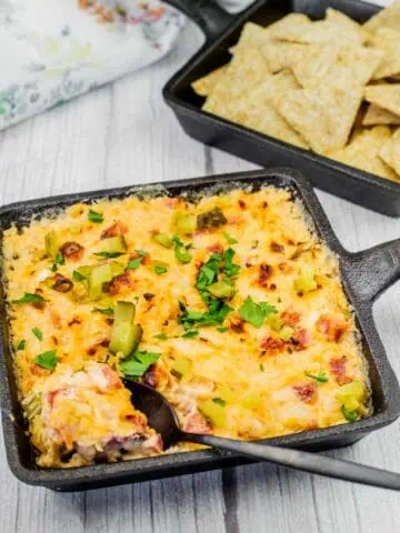 Hot Reuben dip in a cast-iron skillet, garnished with chopped pickles and fresh parsley, served with crispy crackers for dipping.