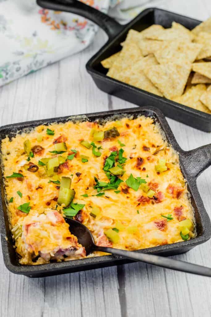Hot Reuben dip in a cast-iron skillet, garnished with chopped pickles and fresh parsley, served with crispy crackers for dipping.
