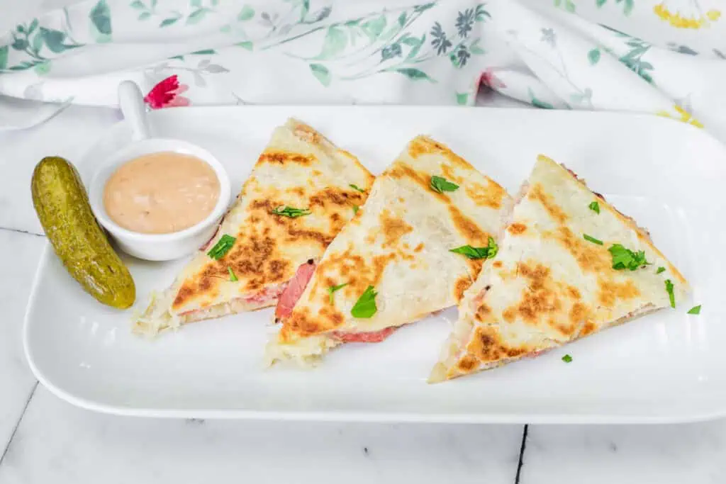 Three quesadilla slices with herbs on a white plate, accompanied by a pickle and a small dish of dipping sauce.