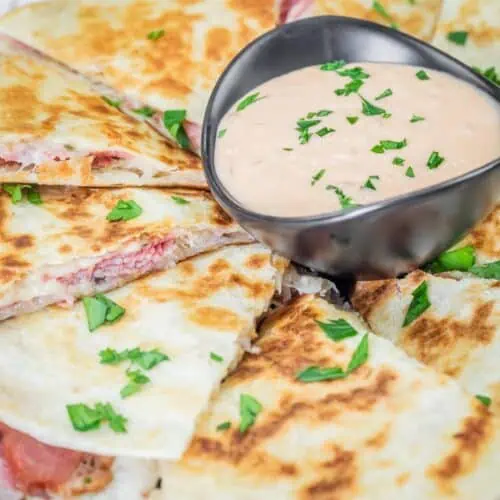Sliced quesadilla with a browned crust, served with a bowl of creamy sauce garnished with chopped herbs on top.