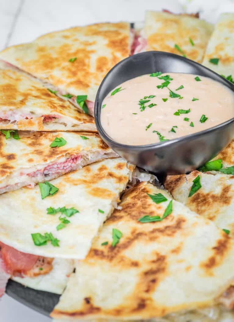 Sliced quesadilla with a browned crust, served with a bowl of creamy sauce garnished with chopped herbs on top.