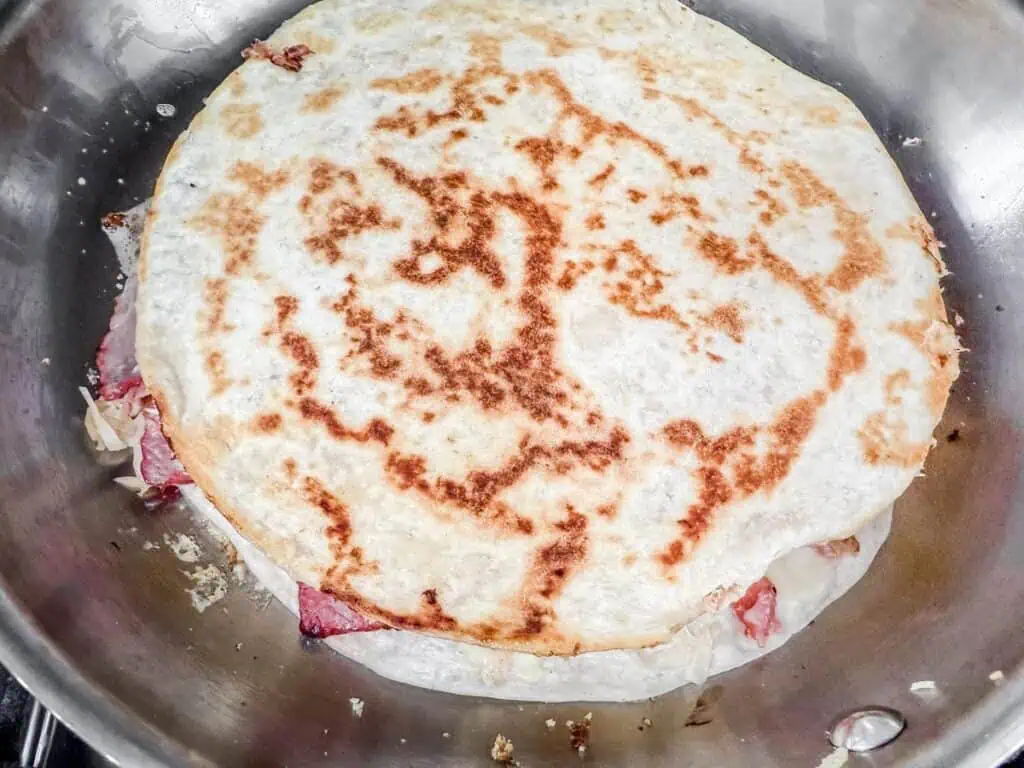 Close-up of a quesadilla with a golden-brown toasted flour tortilla, containing visible melted cheese and meat, cooking in a stainless steel pan.
