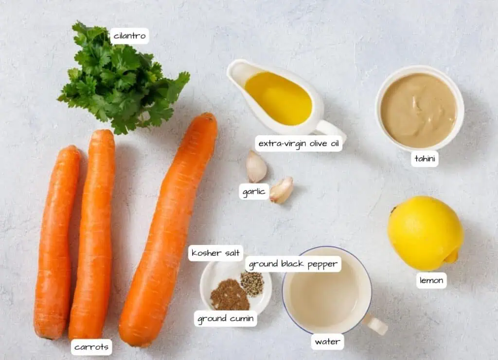 Ingredients on a surface: cilantro, carrots, a lemon, garlic cloves, and bowls of extra-virgin olive oil, tahini, ground cumin, ground black pepper, kosher salt, and water.