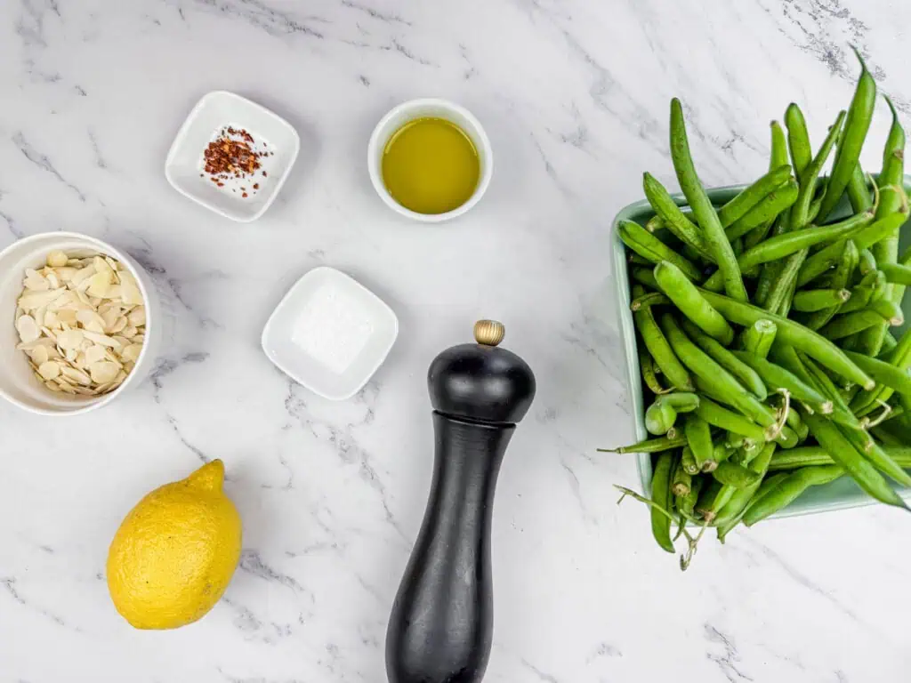 Ingredients arranged on a marble surface: green beans, olive oil, red pepper flakes, sliced almonds, lemon, salt, and a pepper mill.
