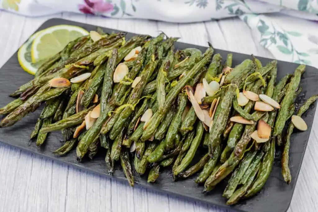Roasted green beans topped with sliced almonds on a black rectangular plate, garnished with lemon slices, on a white wooden surface.