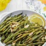 A white dish with roasted green beans topped with sliced almonds and lemon zest, garnished with lemon wedges. Floral decor is visible in the background.