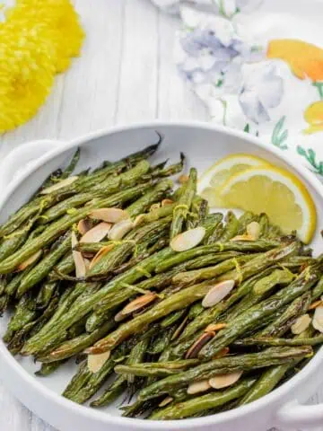 A white dish with roasted green beans topped with sliced almonds and lemon zest, garnished with lemon wedges. Floral decor is visible in the background.