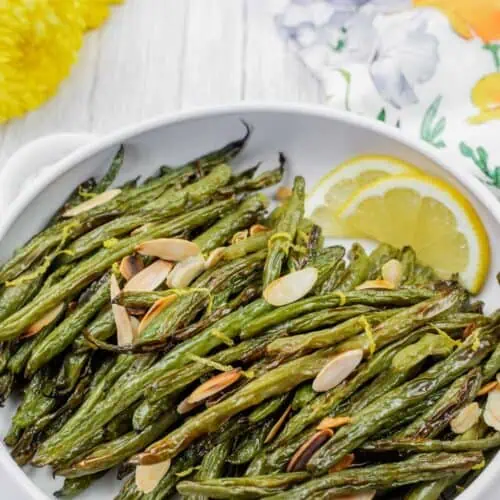 A white dish with roasted green beans topped with sliced almonds and lemon zest, garnished with lemon wedges. Floral decor is visible in the background.