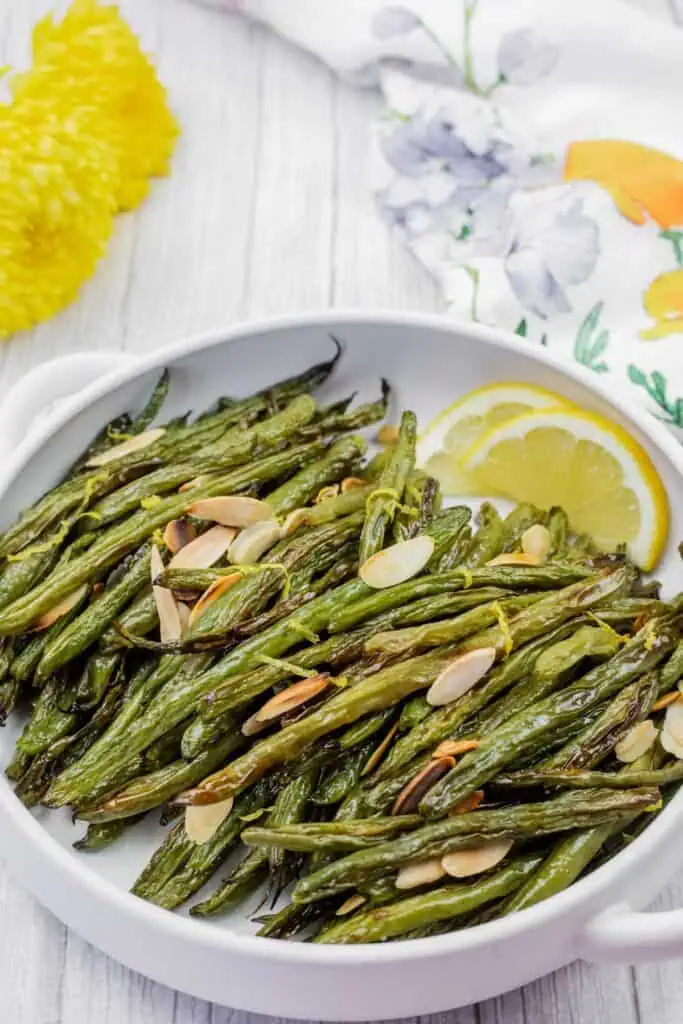 A white dish with roasted green beans topped with sliced almonds and lemon zest, garnished with lemon wedges. Floral decor is visible in the background.