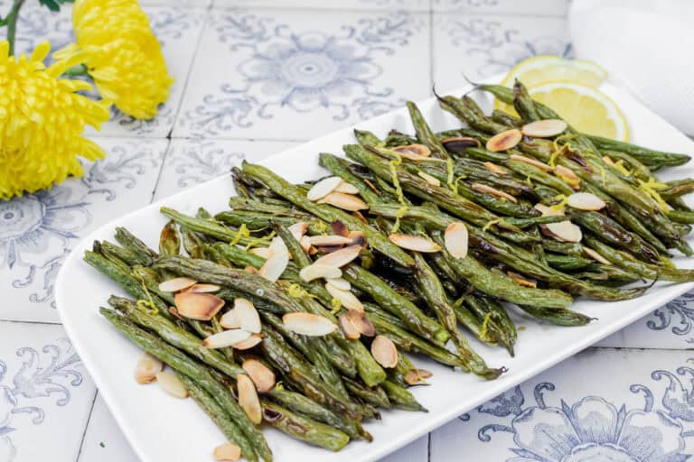 A plate of roasted green beans topped with sliced almonds, garnished with lemon slices on a decorative tiled surface. Yellow flowers are visible in the background.