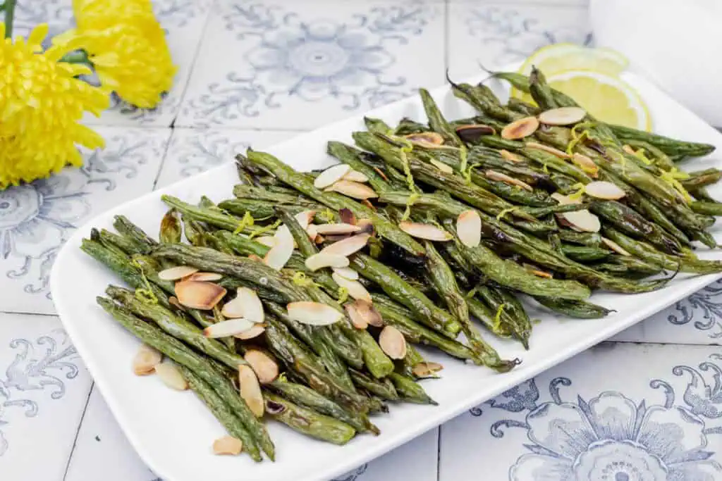 Plate of roasted green beans topped with almond slices and lemon zest, set on a decorative tile surface with yellow flowers and lemon slices in the background.