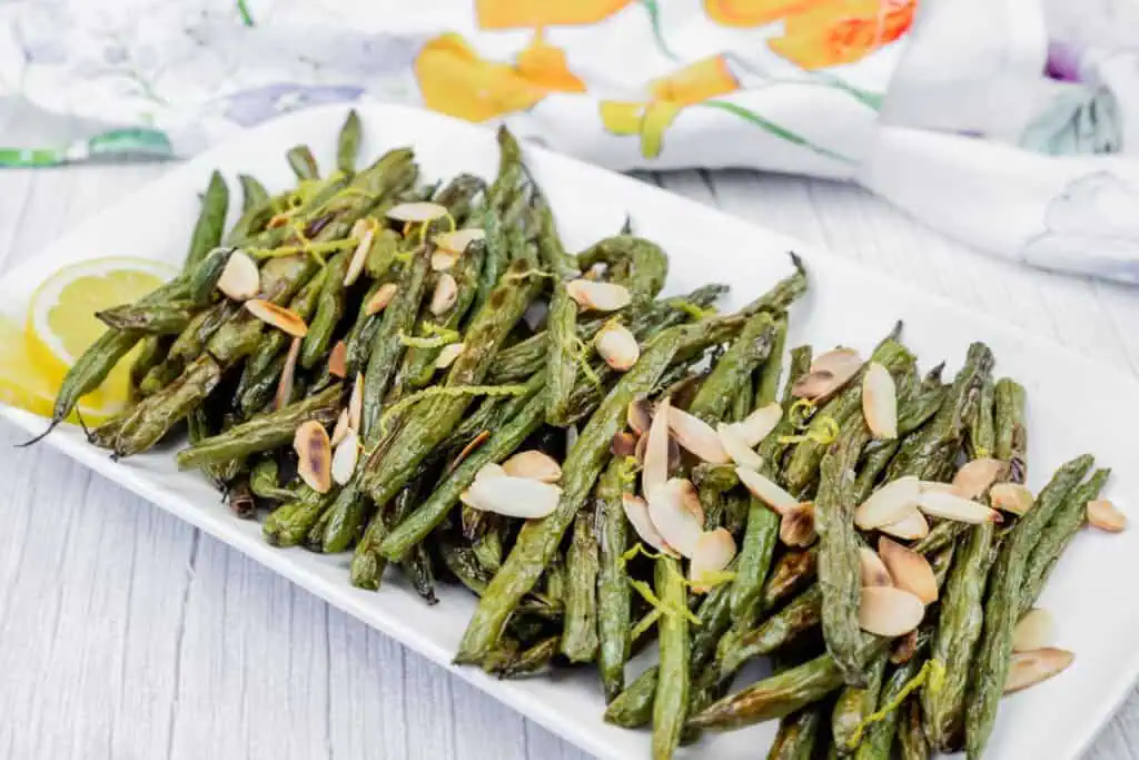 A rectangular white plate with roasted green beans topped with almond slices and lemon zest, garnished with lemon wedges, on a light wooden surface.